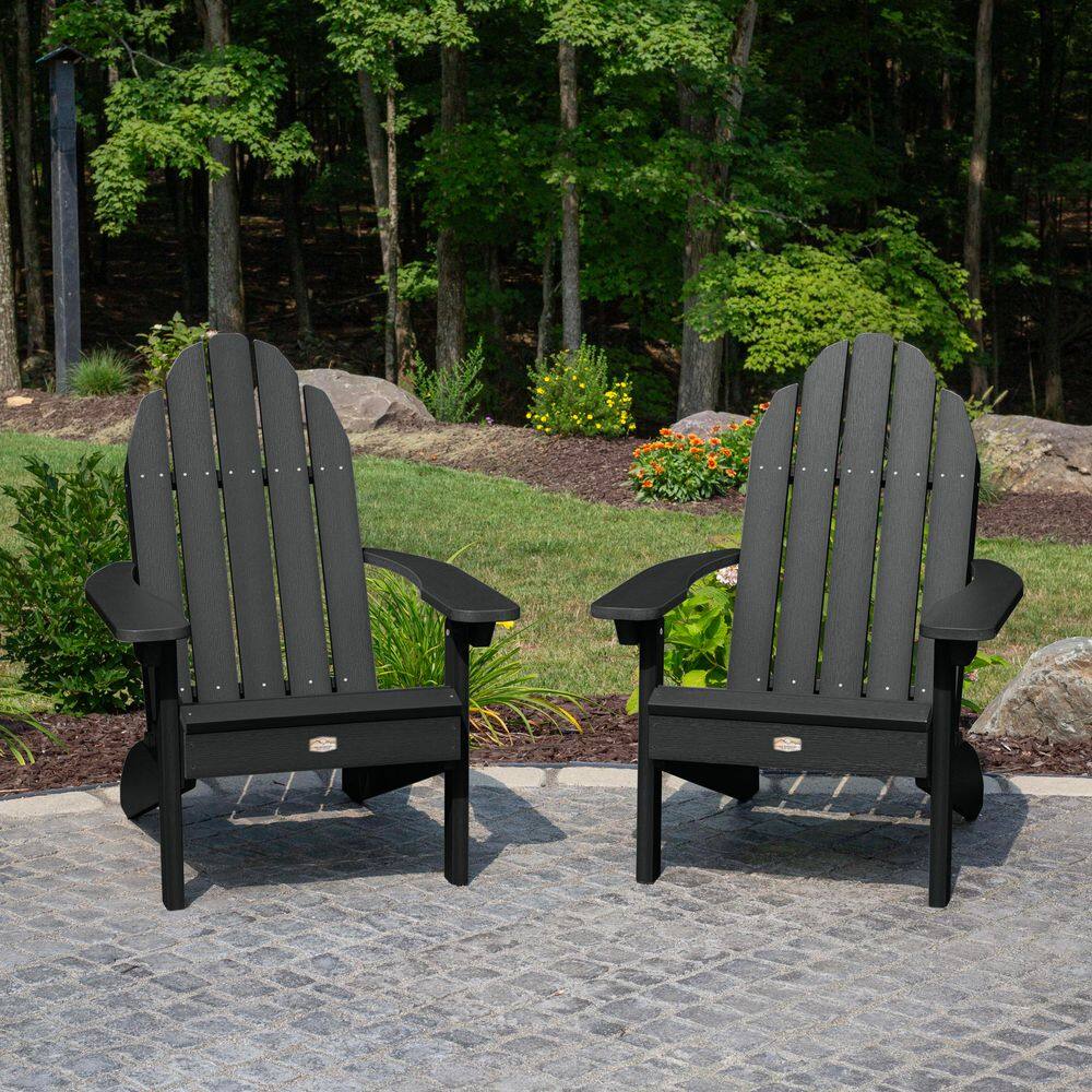 Two black plastic Adirondack chairs on a stone patio in a yard.