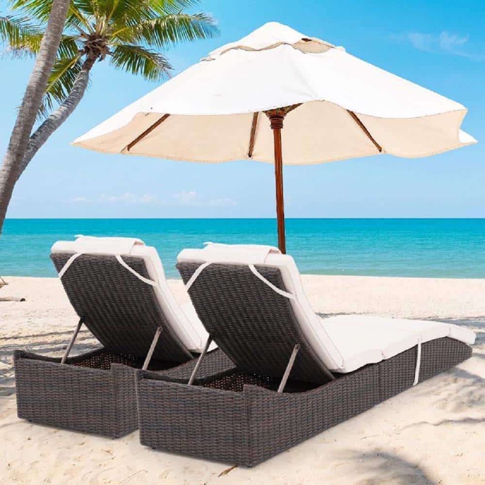 Two brown wicker chaise lounge recliners with beige cushions under a white umbrella on a beach.